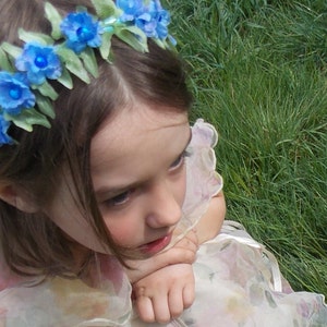 May include: A young girl with long brown hair wears a blue flower crown. She is wearing a white dress with a floral pattern. The girl is sitting in a grassy field.
