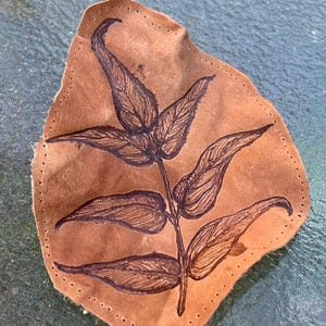 May include: A tan leather patch with an irregular shape featuring a dark brown hand-drawn illustration of a leafy branch. The edges of the leather are perforated with a series of small, evenly spaced holes for stitching, creating a border.