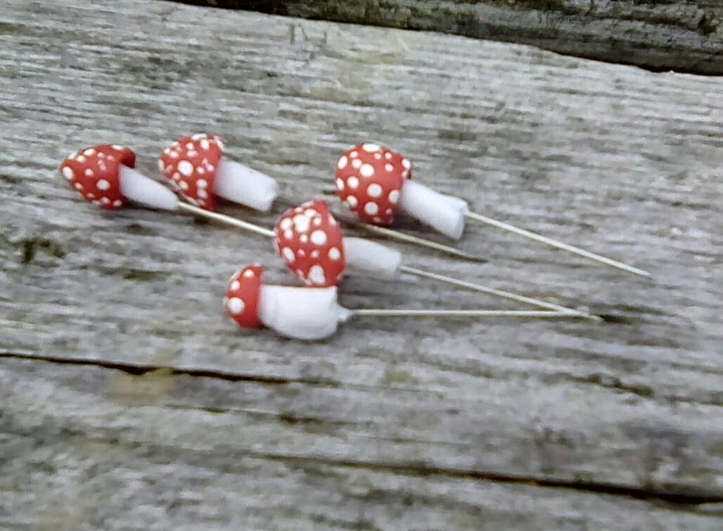 K&ouml;nnte beinhalten: Vier rote und wei&szlig;e Pilz-f&ouml;rmige Stecknadeln mit wei&szlig;en Stielen. Die Stecknadeln sind auf einer Holzoberfl&auml;che angeordnet.