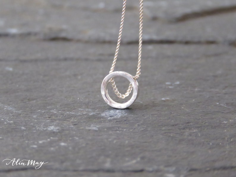 A tiny, hammered circle made of real silver hangs on a delicate sterling silver chain displayed on a black surface.