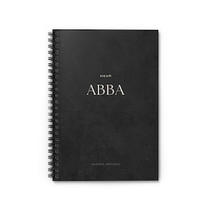 May include: A black spiral-bound notebook with the words "DEAR ABBA" in a serif font. The notebook has a textured cover and the words "NAZARA ARTISAN" are printed at the bottom. The notebook is on a white background.