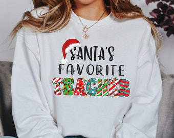 Könnte beinhalten: Weißer Pullover mit einer roten Weihnachtsmütze und dem Text "Santas Lieblingslehrer" in bunten Buchstaben mit Weihnachtslichtern.