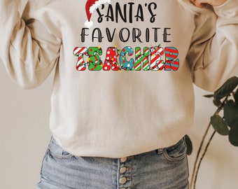 Könnte beinhalten: Ein beiger Pullover mit dem Text "Santa's Favorite Teacher" in bunten Buchstaben mit einer roten Weihnachtsmütze auf dem "S".