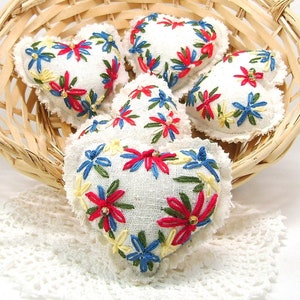 May include: A collection of five heart-shaped ornaments with white fabric and embroidered floral designs in red, blue, and yellow. The ornaments are arranged in a woven basket, with a white lace doily visible beneath.