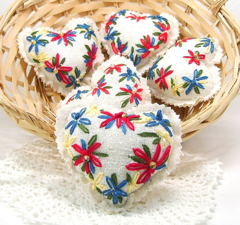 May include: A collection of five heart-shaped ornaments with white fabric and embroidered floral designs in red, blue, and yellow. The ornaments are arranged in a woven basket, with a white lace doily visible beneath.