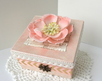 May include: A square, pink decorative box with a large, light pink flower on top. The box features a lace trim, a gold and pink chevron ribbon, and a decorative metal clasp. A piece of sheet music is under the flower.