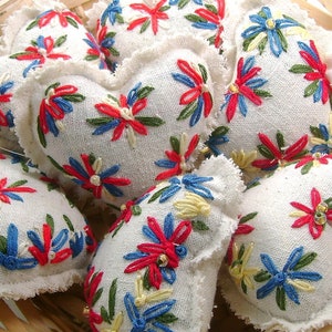 May include: A collection of heart-shaped, embroidered sachets in a woven basket. The sachets are white with colorful floral embroidery in red, blue, green, and yellow. The edges of the sachets have a ruffled trim. The basket is made of light-colored wood.