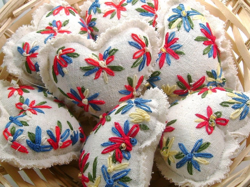 May include: A collection of heart-shaped, embroidered sachets in a woven basket. The sachets are white with colorful floral embroidery in red, blue, green, and yellow. The edges of the sachets have a ruffled trim. The basket is made of light-colored wood.