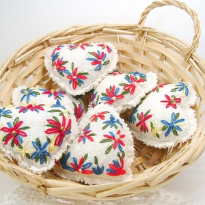 May include: A collection of heart-shaped, embroidered sachets in a woven basket. The sachets are white with colorful floral embroidery in red, blue, and yellow. The basket is made of light-colored woven wood.