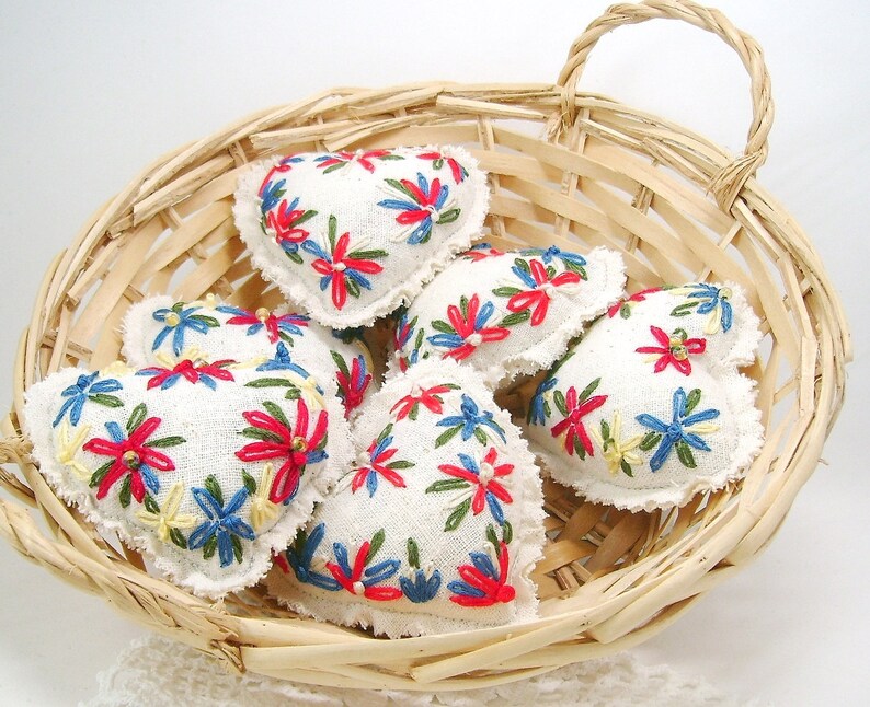 May include: A collection of heart-shaped, embroidered sachets in a woven basket. The sachets are white with colorful floral embroidery in red, blue, and yellow. The basket is made of light-colored woven wood.