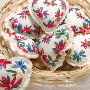 May include: A collection of heart-shaped sachets in a woven basket. Each sachet is white with colorful embroidered floral designs in red, blue, and yellow. The edges of the sachets have a frayed, textured trim. The basket is made of light brown woven material.