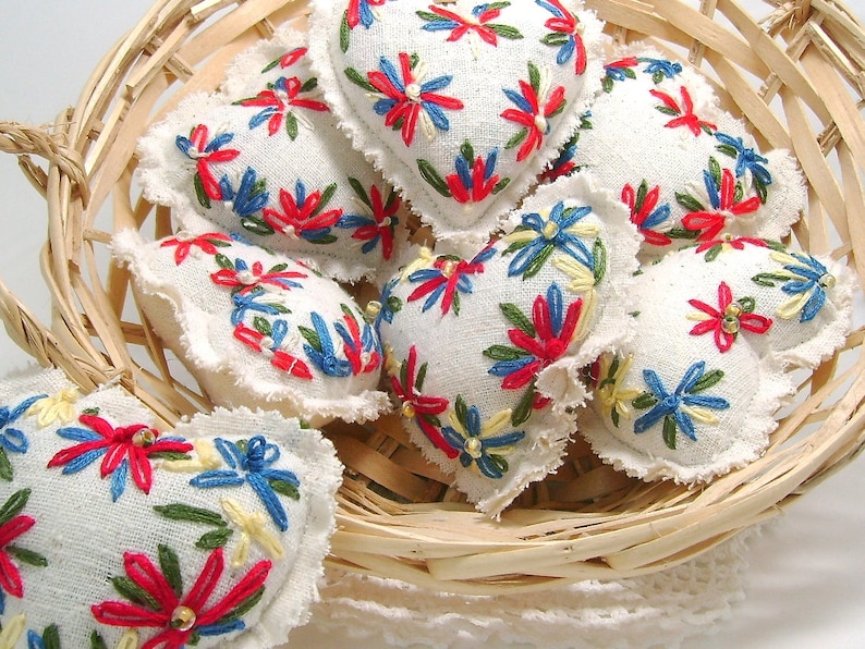 May include: A collection of heart-shaped sachets in a woven basket. Each sachet is white with colorful embroidered floral designs in red, blue, and yellow. The edges of the sachets have a frayed, textured trim. The basket is made of light brown woven material.