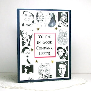 May include: A white card with a blue border features black and white images of famous people. The card has the text "You're in good company, Lefty!" in pink and white.