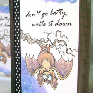 May include: A whimsical notebook with a bat illustration and the text "don't go batty, write it down." The cover features a black ribbon with white polka dots. The bat is brown with large eyes and is hanging upside down.