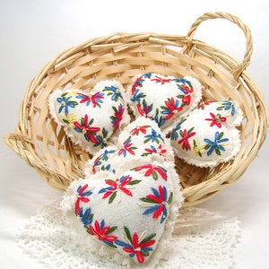 May include: A woven basket holds several white heart-shaped sachets. Each sachet is embroidered with a floral design in red, blue, and yellow. The basket sits on a white lace doily, creating a charming, handcrafted aesthetic.