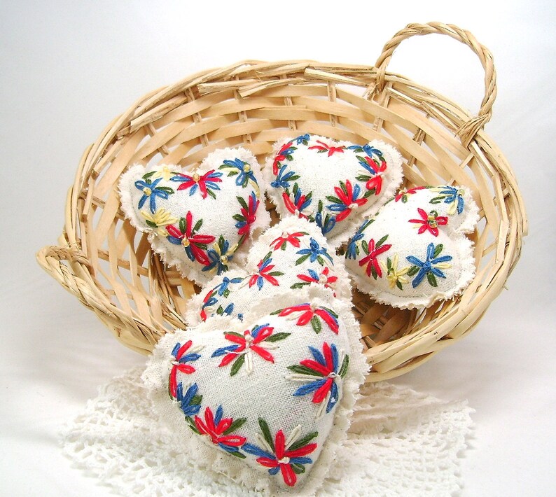 May include: A woven basket holds several white heart-shaped sachets. Each sachet is embroidered with a floral design in red, blue, and yellow. The basket sits on a white lace doily, creating a charming, handcrafted aesthetic.