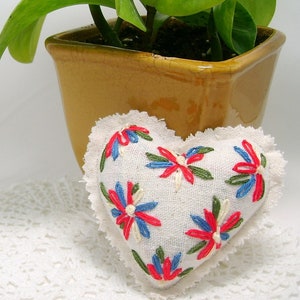 May include: A heart-shaped, off-white fabric pillow with embroidered red, blue, and green floral designs. The pillow has a ruffled edge and is placed on a white, lace-like surface. A yellow-brown ceramic pot with a green plant is in the background.
