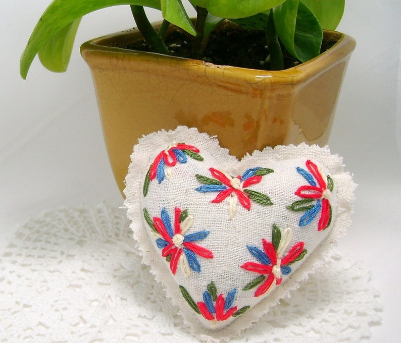 May include: A heart-shaped, off-white fabric pillow with embroidered red, blue, and green floral designs. The pillow has a ruffled edge and is placed on a white, lace-like surface. A yellow-brown ceramic pot with a green plant is in the background.