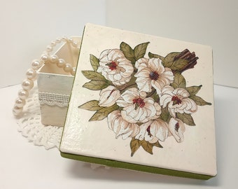 May include: A square, off-white wooden box with a hinged lid, decorated with a floral design featuring white flowers and green leaves. The box has a green velvet trim. A smaller, open box and a pearl necklace are also visible.
