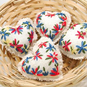May include: Four heart-shaped embroidered sachets with white fabric and colorful floral designs in a woven basket. The embroidery features red, blue, and yellow flowers. The basket is made of light-colored woven wood.