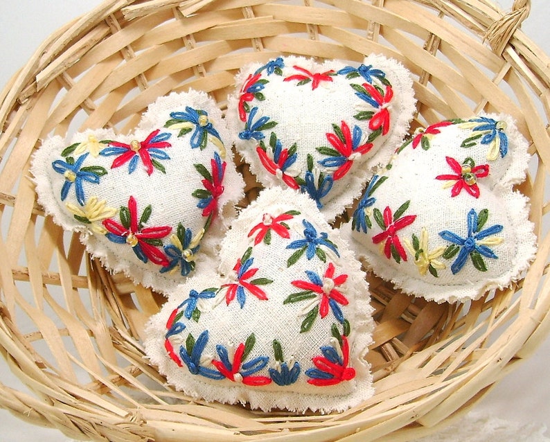 May include: Four heart-shaped embroidered sachets with white fabric and colorful floral designs in a woven basket. The embroidery features red, blue, and yellow flowers. The basket is made of light-colored woven wood.