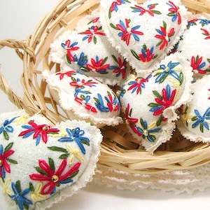 May include: A collection of heart-shaped ornaments with white fabric, embroidered with red, blue, yellow, and green floral designs. The ornaments are in a woven basket, with a white lace doily underneath.