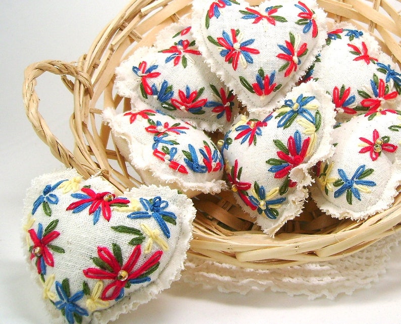 May include: A collection of heart-shaped ornaments with white fabric, embroidered with red, blue, yellow, and green floral designs. The ornaments are in a woven basket, with a white lace doily underneath.