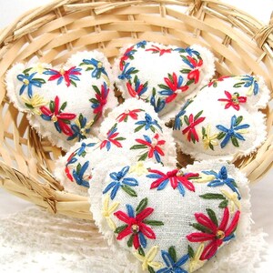 May include: A collection of white heart-shaped sachets with embroidered floral designs in red, blue, and yellow. The sachets are displayed in a woven basket, creating a charming, handcrafted aesthetic. The embroidery details add a touch of color and texture.