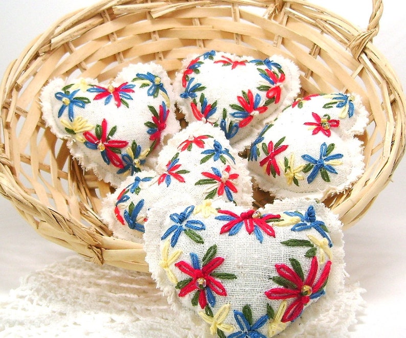 May include: A collection of white heart-shaped sachets with embroidered floral designs in red, blue, and yellow. The sachets are displayed in a woven basket, creating a charming, handcrafted aesthetic. The embroidery details add a touch of color and texture.