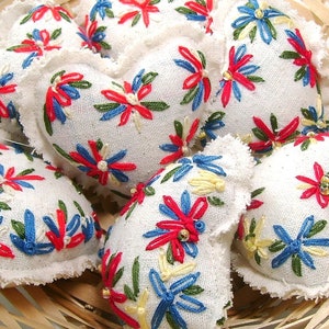 May include: A collection of white fabric heart-shaped ornaments with embroidered floral designs in red, blue, green, and yellow. The ornaments have a frayed edge and are arranged in a woven basket.