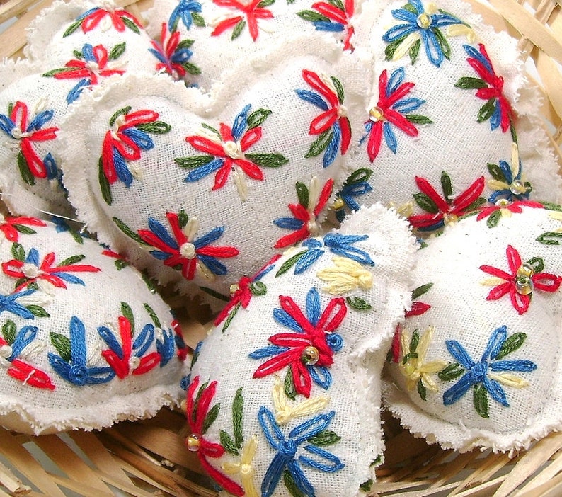 May include: A collection of white fabric heart-shaped ornaments with embroidered floral designs in red, blue, green, and yellow. The ornaments have a frayed edge and are arranged in a woven basket.