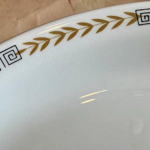 May include: Close-up of a white ceramic plate with a gold and black geometric and leaf design around the rim. The plate has a glossy finish and a simple, elegant aesthetic.