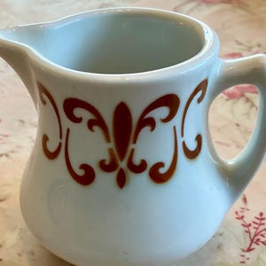 May include: A small, white ceramic creamer with a handle and spout. The creamer features a brown, ornate design around the body. The creamer is likely vintage and suitable for serving cream or milk at a table setting.