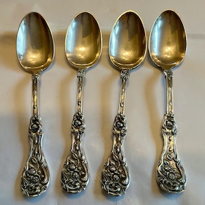 May include: Four silver spoons with ornate floral detailing on the handles. The spoons have a classic design with a rounded bowl and a decorative handle featuring roses and leaves. The spoons are arranged in a row, showcasing their elegant craftsmanship.