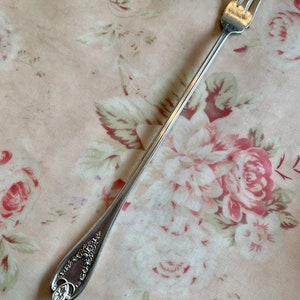 May include: A silver serving fork with three tines, resting on a pink floral patterned surface. The handle features an ornate floral design. This vintage utensil is likely for serving appetizers or desserts.