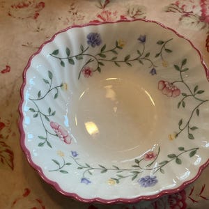 May include: A white ceramic bowl with a pink rim and a floral pattern. The bowl has a scalloped edge and is decorated with small pink, purple, and yellow flowers.
