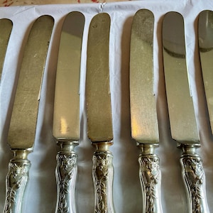 May include: A collection of eight antique butter knives. The blades are a dull silver color, and the handles are ornate with a floral design. The knives are arranged in a row, angled slightly towards the viewer.