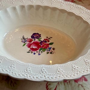 May include: A white, oval-shaped ceramic serving bowl with a scalloped edge and a floral design in the center. The floral design features pink, purple, and blue flowers. The bowl has a decorative, embossed border.