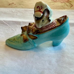 May include: A whimsical ceramic figurine featuring a mouse wearing a chef's hat and checkered scarf, nestled inside a turquoise high-heeled shoe. The shoe has decorative accents and a glossy finish, perfect for a collector.
