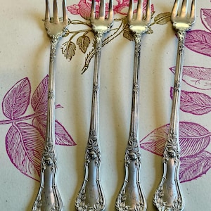 May include: Four antique silver forks with ornate floral detailing. Each fork has a slender handle with a rose design at the base and a three-tine head. The forks are arranged on a floral patterned background.