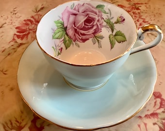 May include: A light blue porcelain teacup and saucer set featuring a gold-trimmed rim and handle. The interior of the cup displays a detailed illustration of a pink rose with green leaves and stems, while the saucer has a matching gold edge.