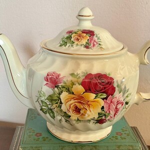 May include: Cream-colored porcelain teapot with a lid, adorned with a floral rose pattern in shades of red, yellow, and pink. The teapot features a curved spout, a loop handle, and gold trim accents. A decorative piece for serving tea.