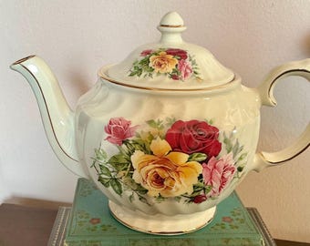 May include: Cream-colored porcelain teapot with a lid, adorned with a floral rose pattern in shades of red, yellow, and pink. The teapot features a curved spout, a loop handle, and gold trim accents. A decorative piece for serving tea.