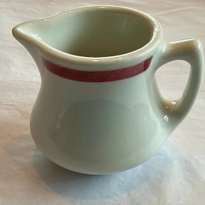 May include: A small, off-white ceramic creamer with a red stripe around the top. The creamer has a curved body, a handle, and a spout for easy pouring. The creamer is likely vintage or antique.