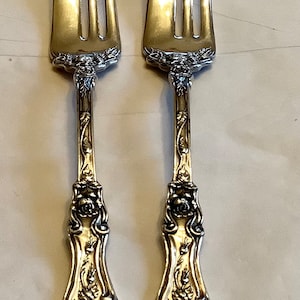 May include: Two silver forks with ornate handles featuring floral designs. The forks have three tines each and are polished to a shine.