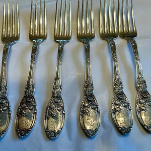 May include: Six silver forks with ornate floral and scroll detailing. The forks have four tines and a decorative handle with an engraved oval cartouche. The silverware is arranged on a white surface, showcasing the intricate design.