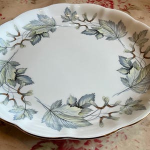 May include: A white porcelain serving platter with a scalloped edge and gold trim. The center features a decorative border of blue and green maple leaves and winged seeds arranged in a circular pattern around the white basin of the dish.