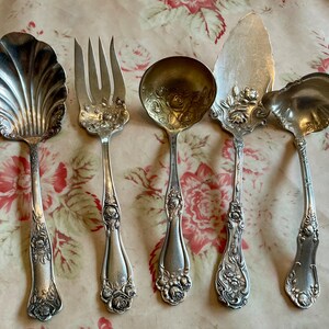 May include: An assortment of antique silver serving utensils, including a shell-shaped spoon, a fork, a round spoon, a cake server, and a ladle. Each piece features ornate floral detailing on the handles and decorative elements.