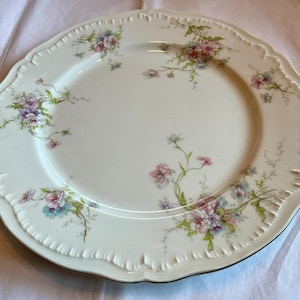 May include: A white porcelain plate with a scalloped edge and floral design. The plate features a central area and a decorative border. The floral pattern includes pink, purple, and blue flowers with green stems and leaves. The plate has a gold trim.