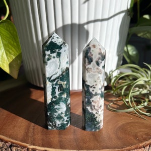 May include: Two green, white, and brown moss agate crystal points on a wooden surface. The points are polished and have a natural, organic look.